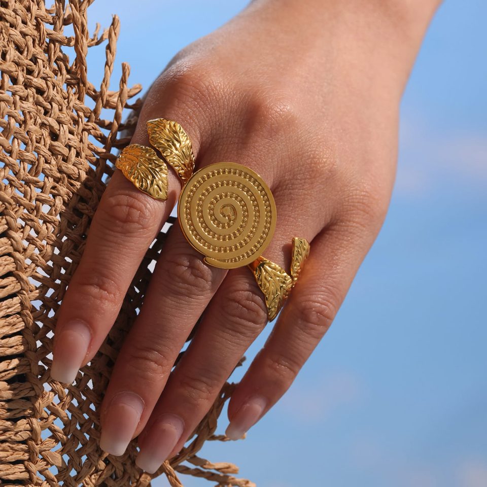 A model wearing a gold circular groove open ring with a hat design, holding a sun hat in front of a blue sky and sea.