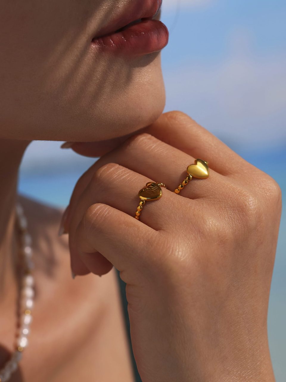 Close-up of model’s hand with gold open heart ring resting on chin, showcasing ring details