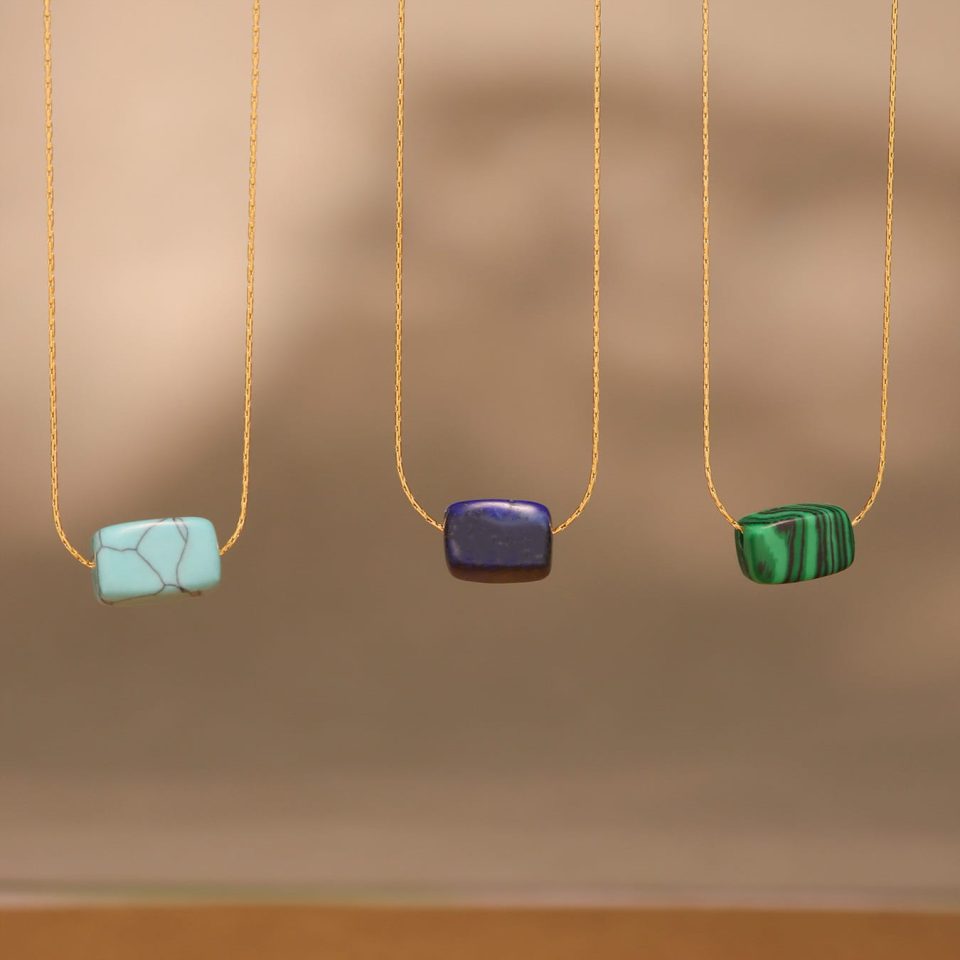 Flat lay of OUMIAKA’s lapis lazuli, blue turquoise, and malachite rectangular pendant necklaces on rustic background, 18K gold-plated chains