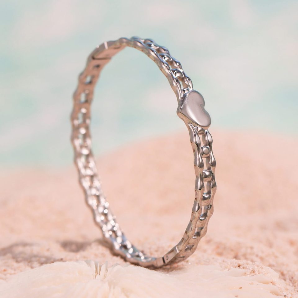 Silver cuff bangle with heart-lock clasp resting on smooth ocean stones